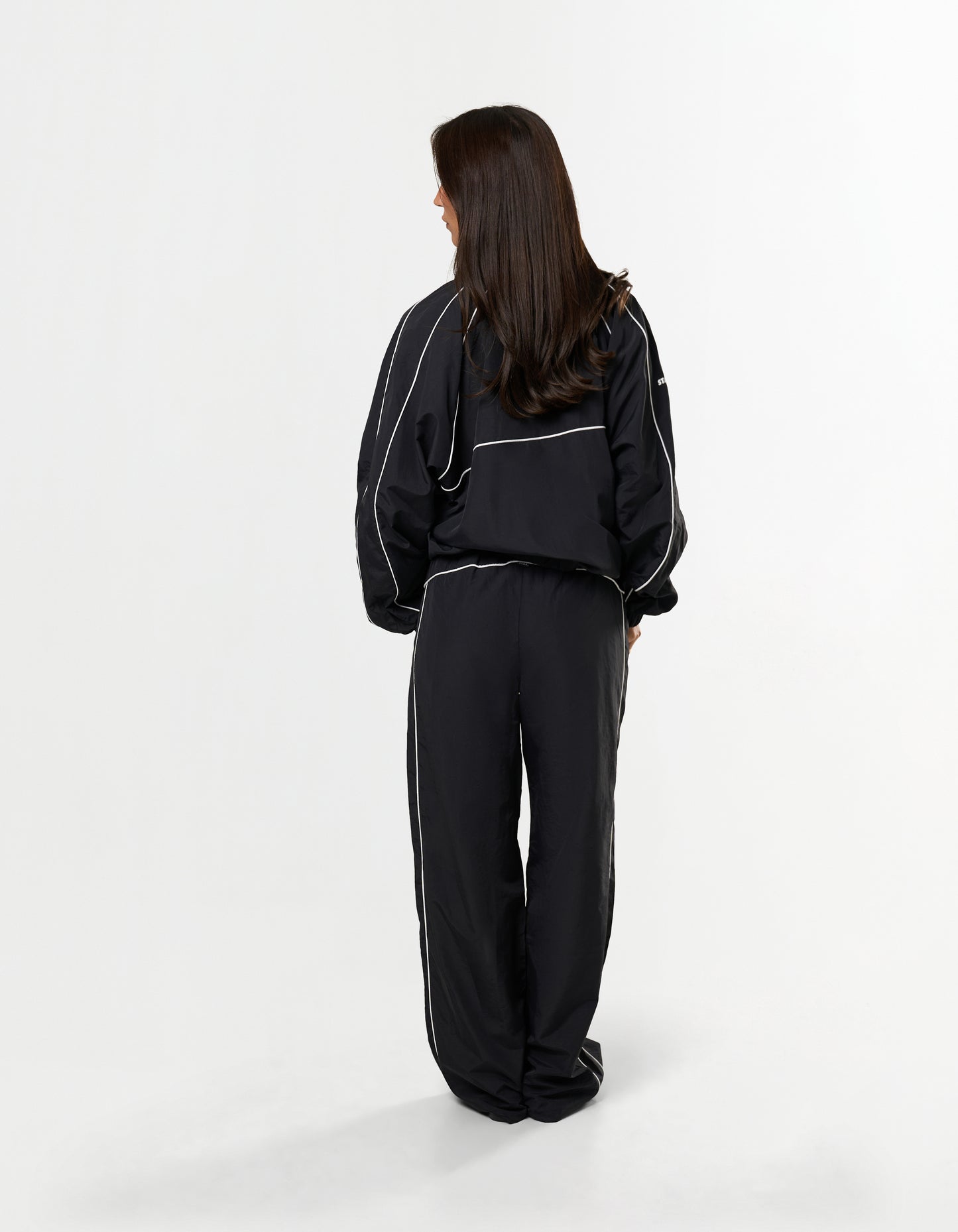 Back view of model wearing STAX. Woven Track Jacket in black with matching Track Pants