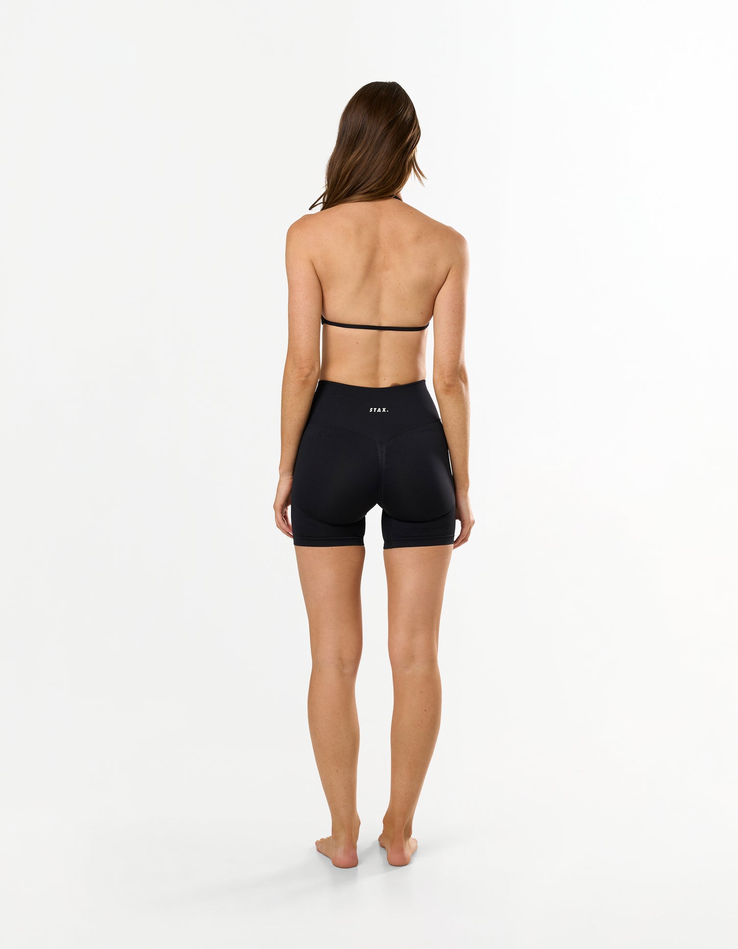 Back view of model wearing STAX. Premium Seamless Skulpt Midi Bike Shorts   in Black showing flattering invisible scrunch detail and matching strappy halter neck bralette